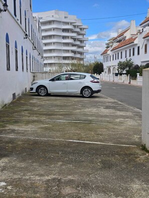 En hvid bil parkeret på en belagt plads ved siden af en hvid bygning med flere balkoner.