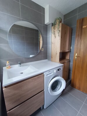 A modern bathroom with a white sink, a built-in washing machine, a wooden cabinet, and a mirror. - Vrbo Property (Geispolsheim)