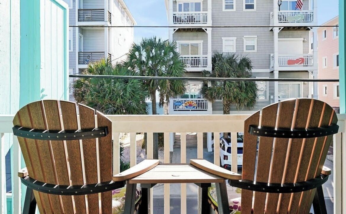 A balcony with two wooden chairs and a small table, overlooking a street with buildings and palm trees.