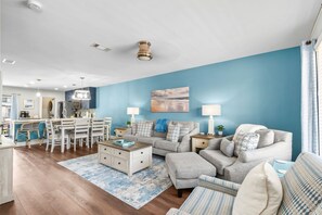 A modern living room with a blue accent wall, a dining area with a white table and chairs, and a kitchen with dark cabinets. - Walkable to Beach, Family Friendly (Miramar Beach)