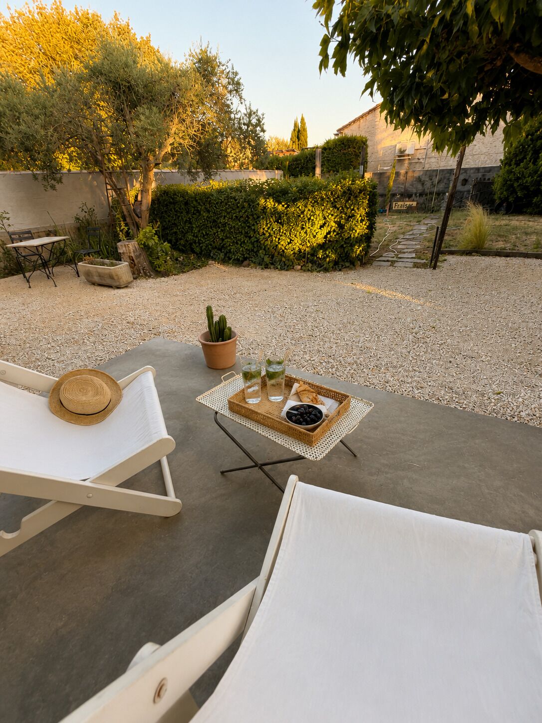 A patio with two white lounge chairs, a small table with a tray of drinks and olives, and a potted cactus.