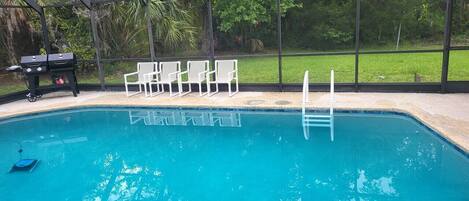 A swimming pool with a glass enclosure, surrounded by chairs and a barbecue grill.