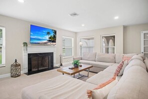 A living room with a fireplace, a large flat-screen TV mounted above it, a sofa, and a coffee table. - Gem at Crystal River (Crystal River)