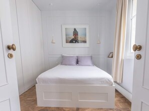 Ein Schlafzimmer mit einem weißen Bett, zwei Kissen, einem gerahmten Bild an der Wand und einem Fenster mit Vorhängen.