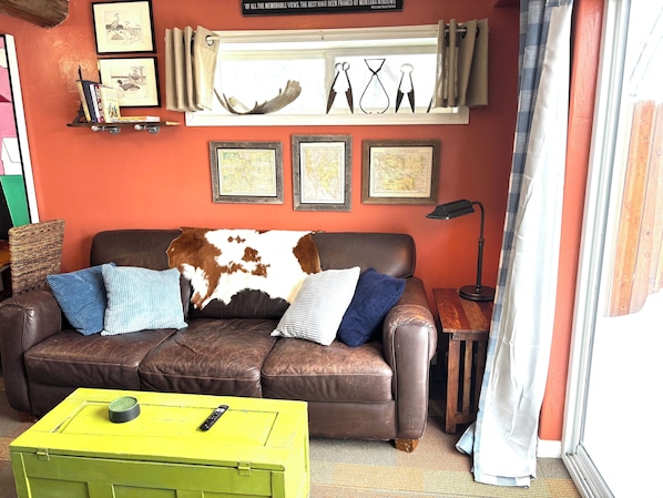 A living room with a brown leather sofa, two blue pillows, a yellow side table with a remote control, and framed maps on the wall.