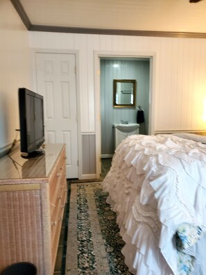 Ein Schlafzimmer mit Bett, Kommode, Fernseher, einer Tür, einem Spiegel und einem durch eine offene Tür einsehbaren Badezimmer.