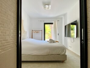 A bedroom with a bed, a TV mounted on the wall, a door leading outside, and a brick wall. - Welcome to Le Jardin des Cambrettes, a 2-hectare private estate at the gate (Lattes)