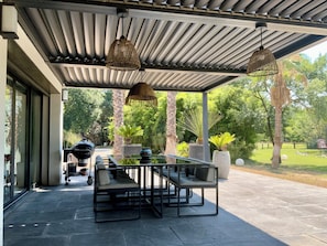 An outdoor patio with a glass table and chairs, surrounded by greenery and a barbecue grill. - Welcome to Le Jardin des Cambrettes, a 2-hectare private estate at the gate (Lattes)