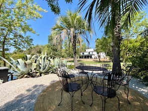 A patio with a round table and chairs, surrounded by large plants and a palm tree, with a building in the background. - Welcome to Le Jardin des Cambrettes, a 2-hectare private estate at the gate (Lattes)