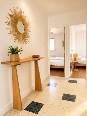 Ein modernes Hotelzimmer mit einem spiegelnden, sonnenförmigen Spiegel, einem Holzkonsolentisch mit Pflanze und Schale, einem Bett mit weißer Bettwäsche und Blick in einen anderen Raum durch eine geöffnete Tür.