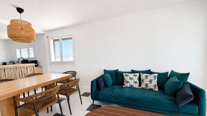 Un soggiorno moderno con un tavolo da pranzo in legno, sedie in vimini, un divano color petrolio con cuscini decorati e una lampada a sospensione.