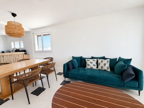 Ein modernes Wohnzimmer mit einem Holzesstisch, Korbstühlen, einem türkisfarbenen Sofa mit gemusterten Kissen und einer Pendelleuchte.
