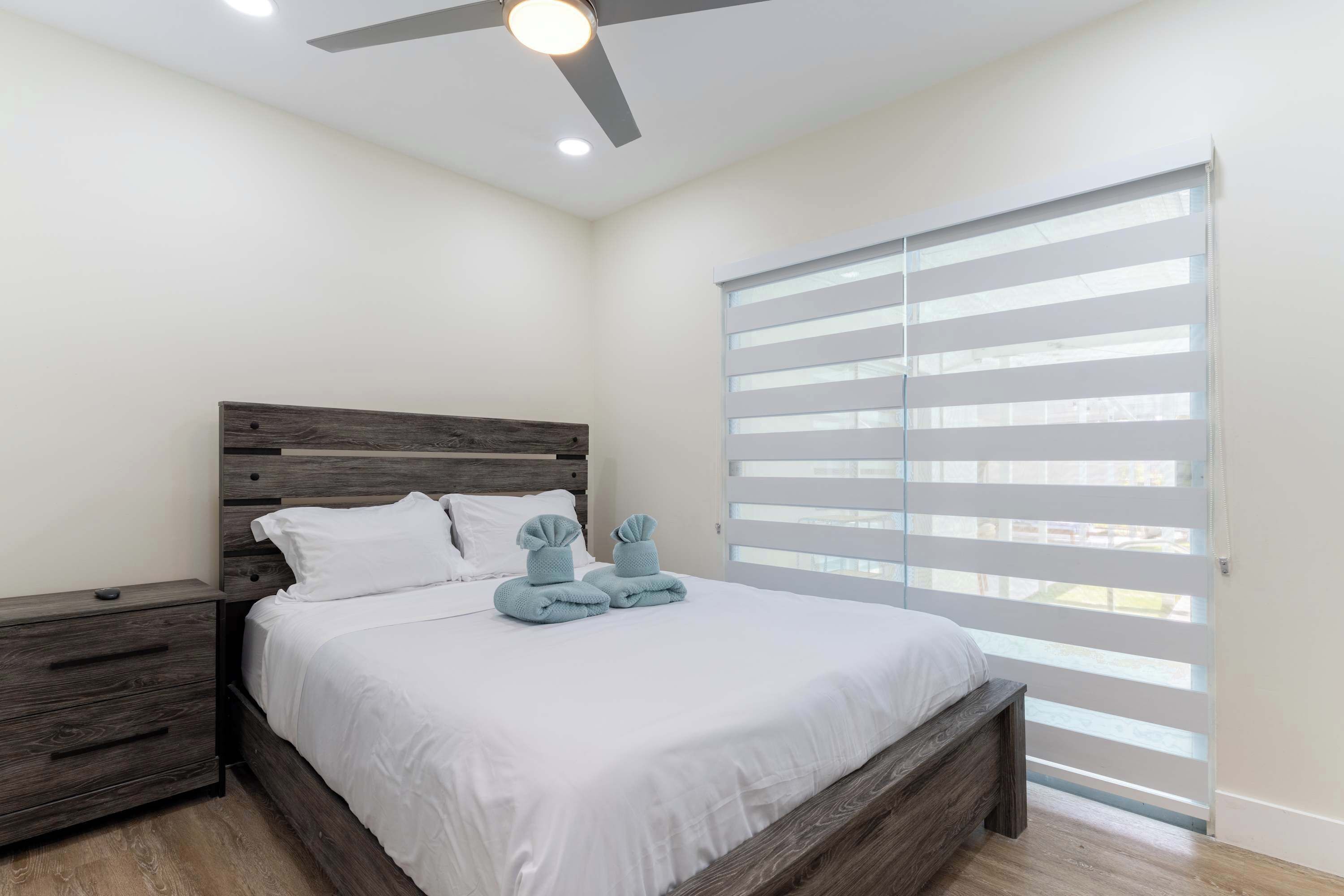 Un dormitorio con cama, mesitas de noche, ventilador de techo y un ventanal con persianas.