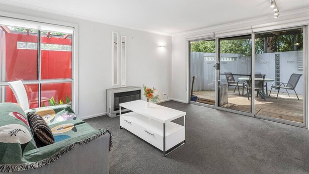 Uma sala de estar moderna com um sofá branco, uma mesa de centro branca e uma porta de vidro deslizante que dá para um pátio externo com mesa e cadeiras.