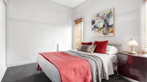 Ein Schlafzimmer mit Bett, Nachttisch, Lampe, einem Gemälde und einem Fenster mit Jalousien.