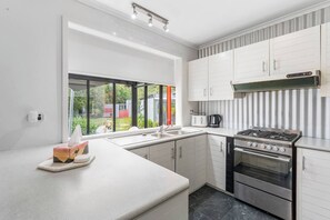 A modern kitchen with white cabinetry, a gas stove, an oven, and a large window offering a view of the outdoors. - Greenview Bungalow Sunlit Stay near Sovereign Hill (Brown Hill)