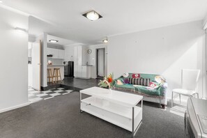 A modern living room with a white sofa, a coffee table, and a kitchen in the background. - Greenview Bungalow Sunlit Stay near Sovereign Hill (Brown Hill)