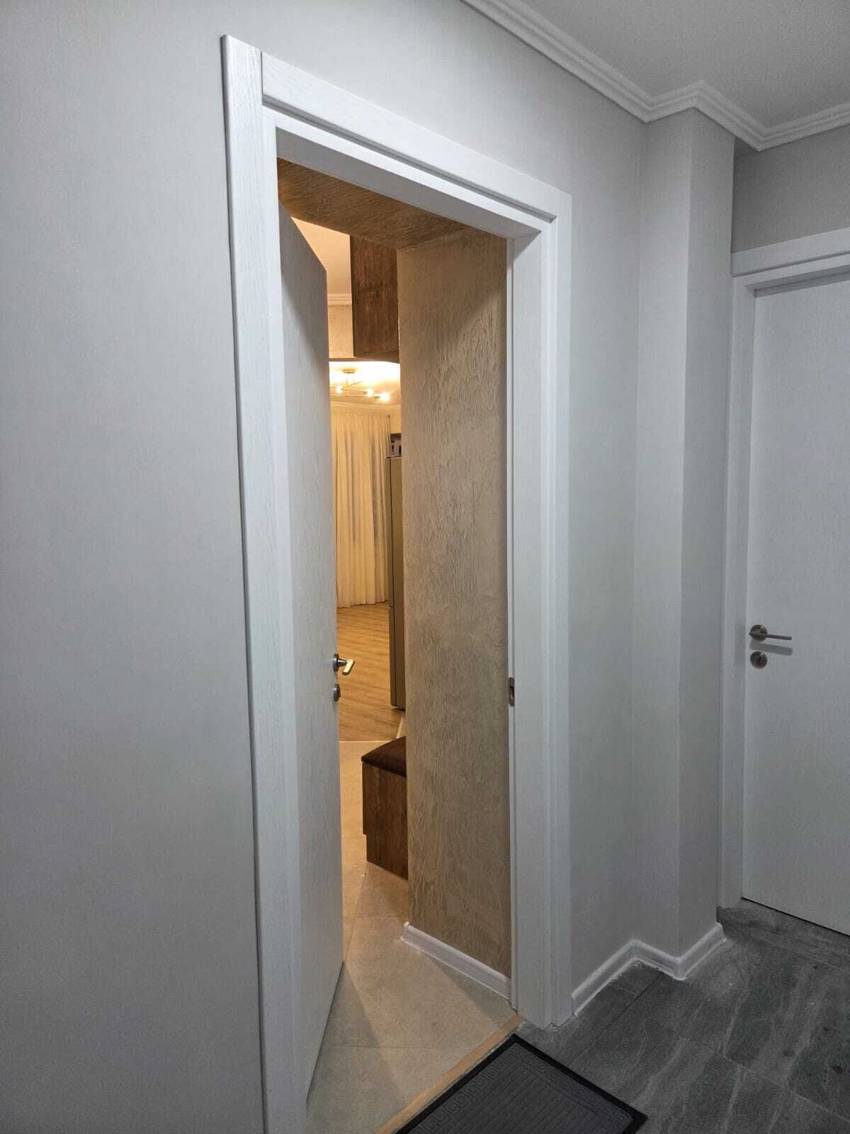 Un couloir avec une porte donnant sur une pièce dont le sol est en bois et qui est équipée d’un luminaire.