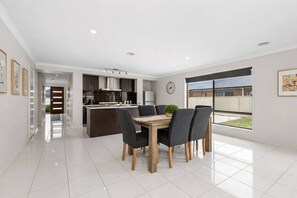 A modern kitchen with dark cabinetry, a central island, and a dining area with a wooden table and chairs. - Chifley Place Modern Comfort near Delacombe Town (Delacombe)