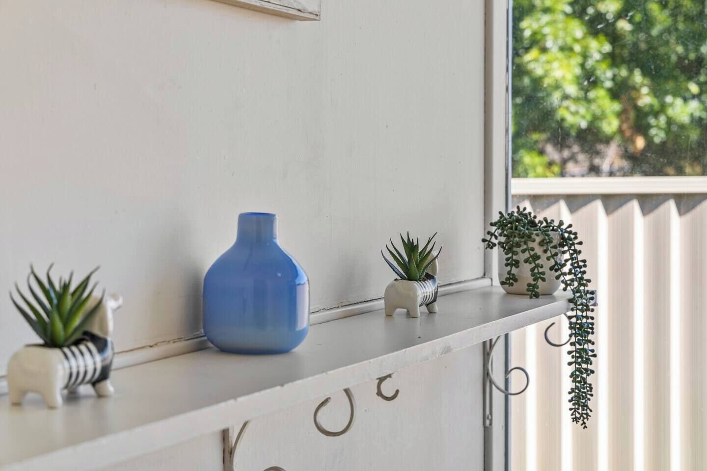 Uno scaffale bianco con un vaso blu, una piccola pianta in vaso e una pianta pensile con gancio metallico.