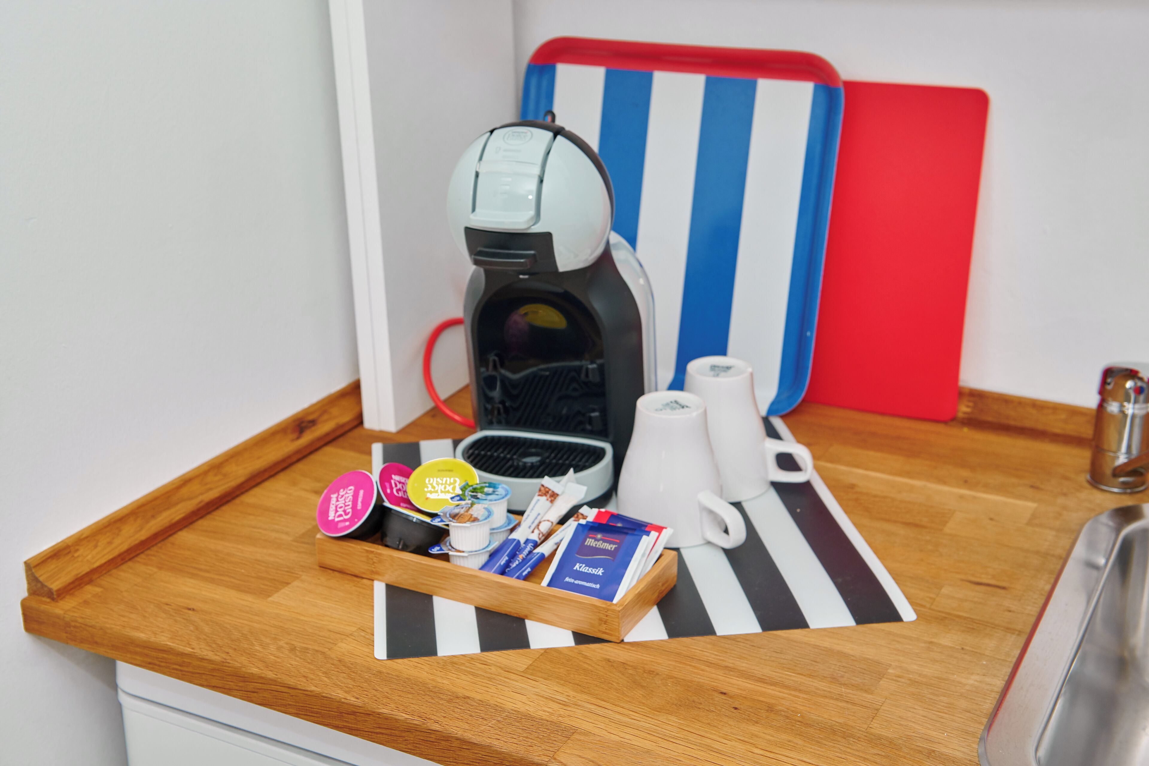 Un angolo cottura con una macchina del caffè, un vassoio con cialde per il caffè, una tazza e un tagliere a righe rosse e blu.