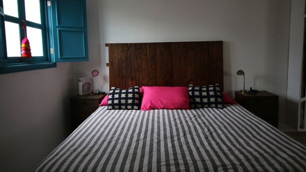 Un dormitorio con cama, ventana con persianas azules, mesita de noche con lámpara y calentador de pared.