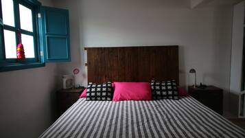 Une chambre à coucher avec un lit, une fenêtre avec des volets bleus, une table de chevet avec une lampe et un radiateur fixé au mur.