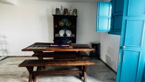 Una habitación con una mesa de madera y bancos, una puerta azul y una estantería con vajilla y frascos.
