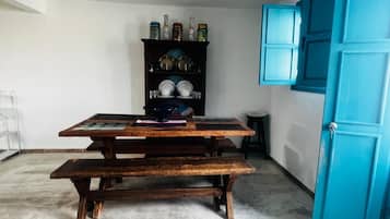 Une pièce avec une table et des bancs en bois, une porte bleue et une étagère où sont posés des assiettes et des bocaux.