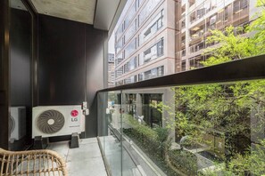 Varanda moderna com guarda-corpo de vidro, com vista para um jardim e edifícios vizinhos.