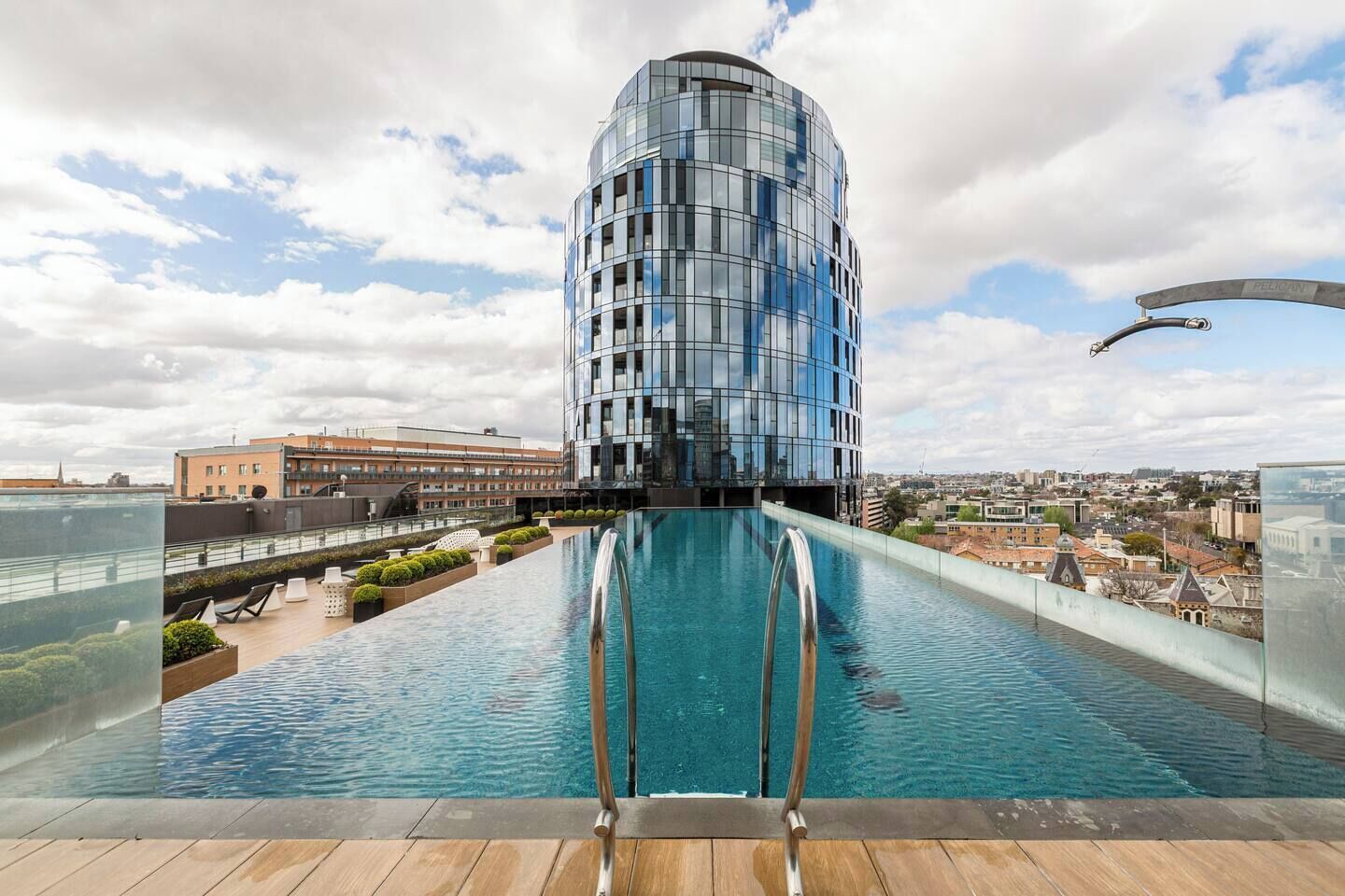 A modern building with a reflective glass facade, a rooftop pool with a wooden deck, and a cityscape in the background.