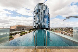 A modern building with a reflective glass facade, a rooftop pool with a wooden deck, and a cityscape in the background. - Parque Canopy Lakeside Living with Infinity Pool (Melbourne)
