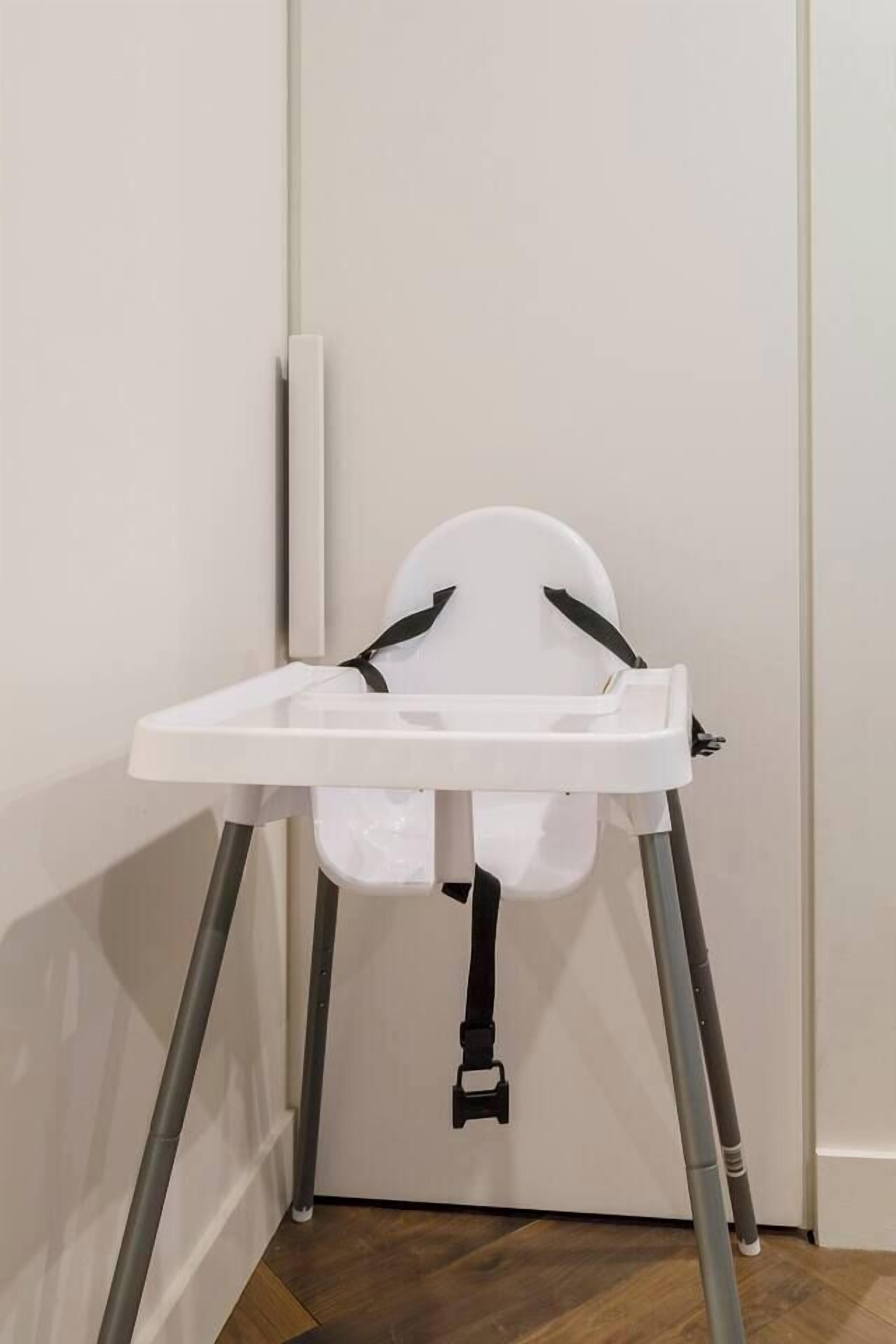 A white baby high chair with a tray, positioned against a wall with a door.