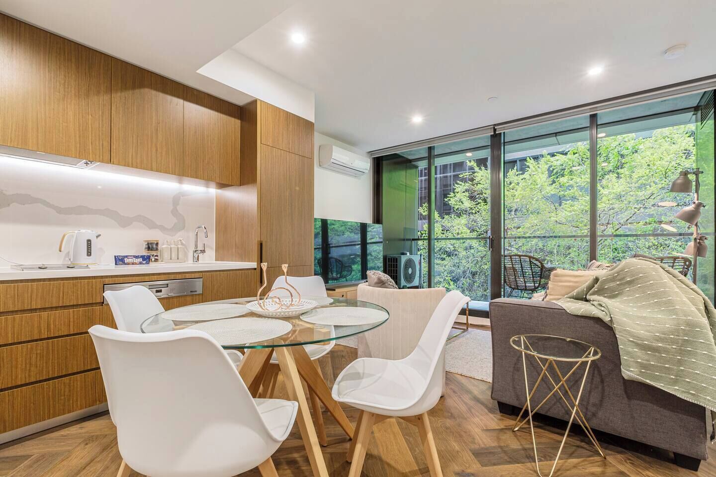 A modern kitchen with a glass dining table and white chairs, a grey sofa, and a view of greenery through large windows.