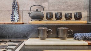 Une cuisine équipée d’une cuisinière à gaz, d’une bouilloire et d’un ensemble de quatre tasses de couleur sombre, posées sur une étagère en bois.