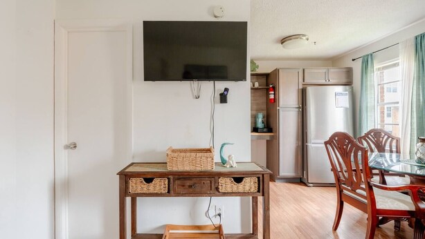 A dining area with a wooden table and chairs, a television mounted on the wall, and a kitchen in the background. - Budget-Friendly 2BR/2BA • Walkable to Events (Virginia Beach)