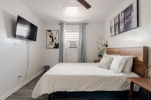 A bedroom with a bed, a TV mounted on the wall, a window with curtains, and a nightstand with a lamp. - ViBe District 2BR/2BA On 19th Street (Virginia Beach)
