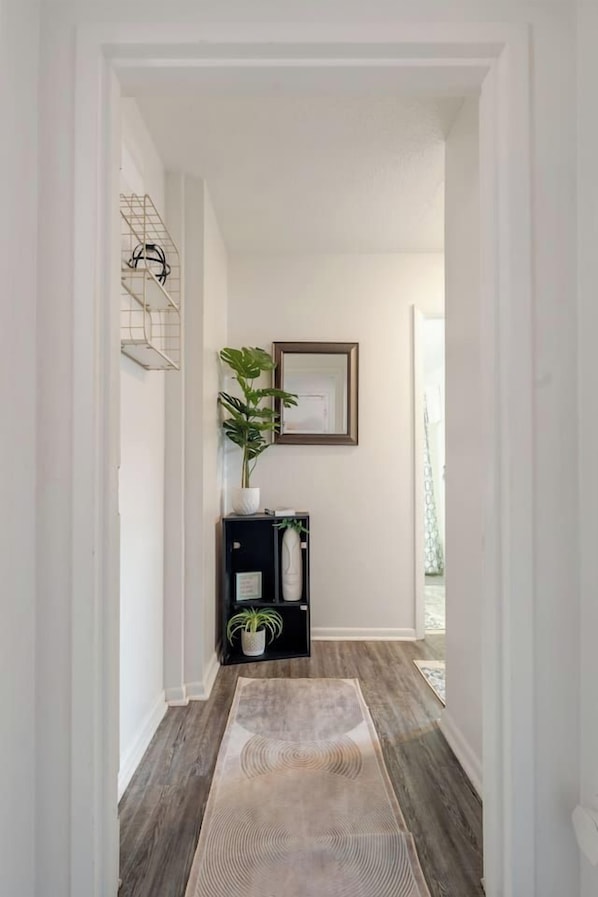 A hallway with a wooden floor, a rug, a plant, a framed picture, and a black cabinet with a plant. - ViBe District 2BR/2BA On 19th Street (Virginia Beach)