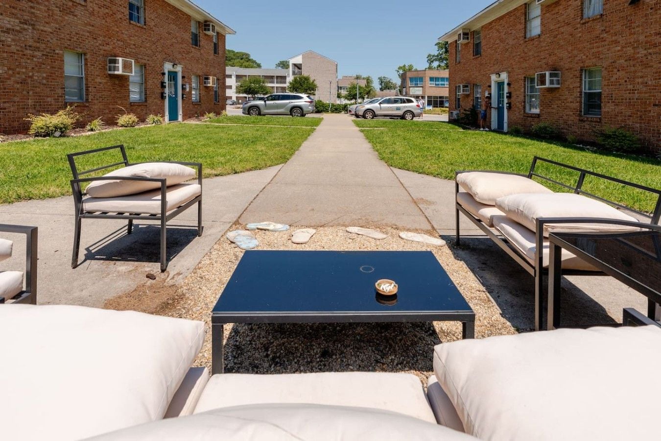 An outdoor seating area with a black table and white cushioned chairs, a gravel path, and brick buildings in the background.