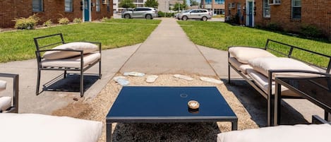 An outdoor seating area with a black table and white cushioned chairs, a gravel path, and brick buildings in the background.