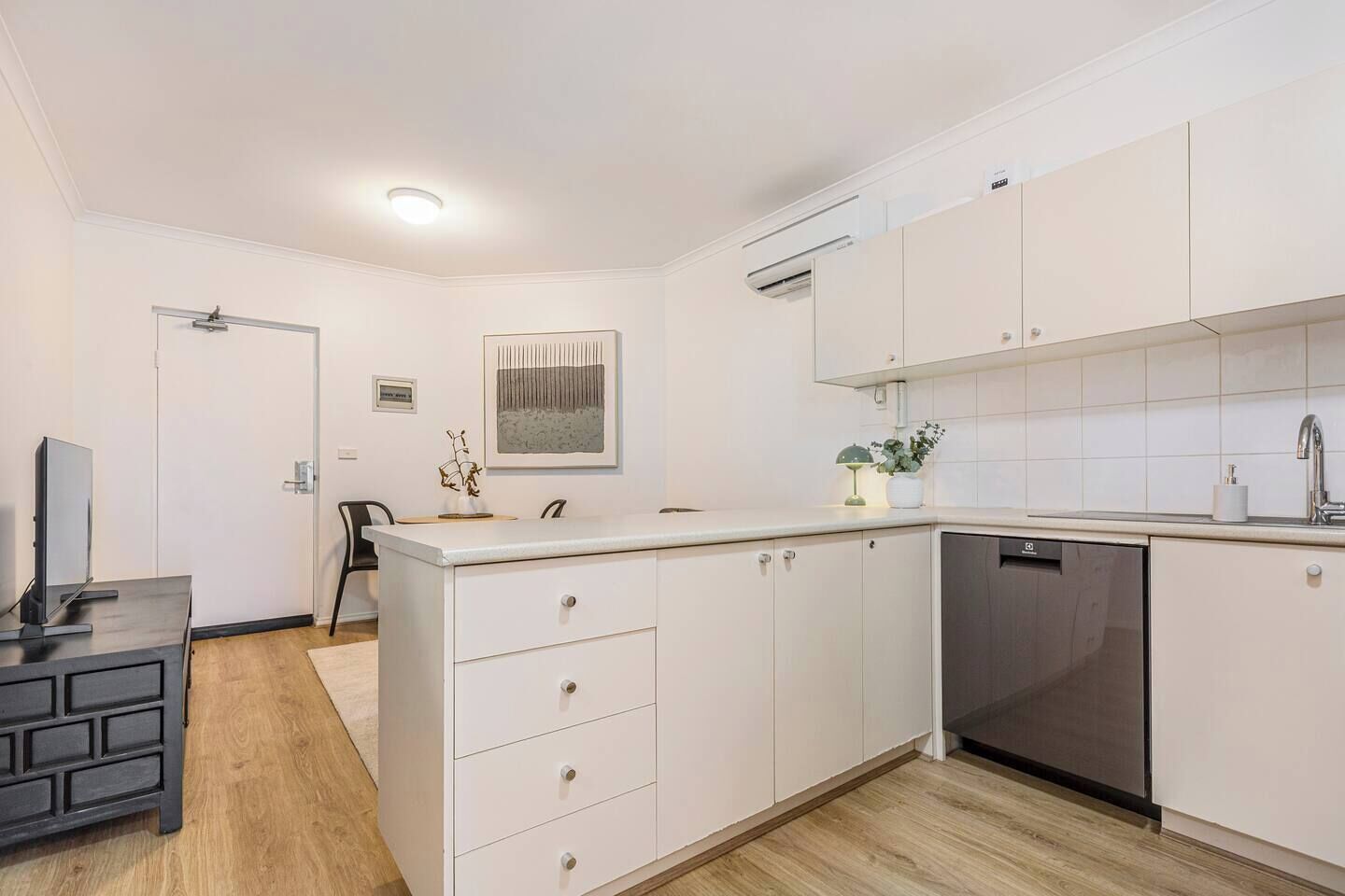 A modern kitchen with white cabinetry, a dishwasher, and a dining area with a TV and a chair.