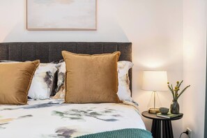 Ein ordentlich bezogenes Bett mit einer blumigen Tagesdecke und zwei Kissen, ein Nachttisch mit einer Lampe und einem Blumenstrauß, sowie ein gerahmtes Bild an der Wand.