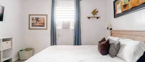 A neatly arranged bedroom with a bed, a framed picture, a shelf with a decorative item, and a window with curtains.