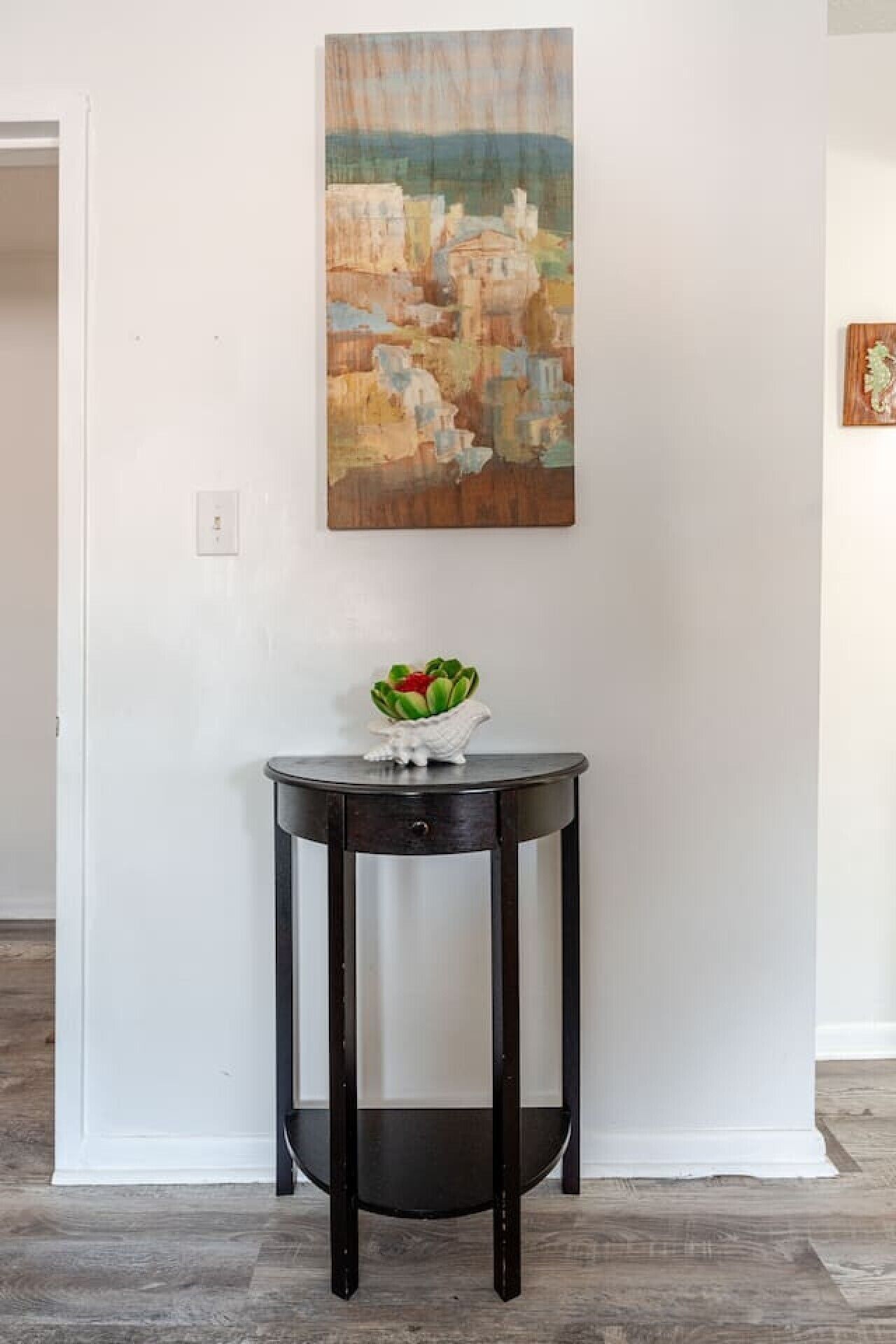 A room with a dark wooden side table against a white wall, a painting above it, and a small white plant on the table.
