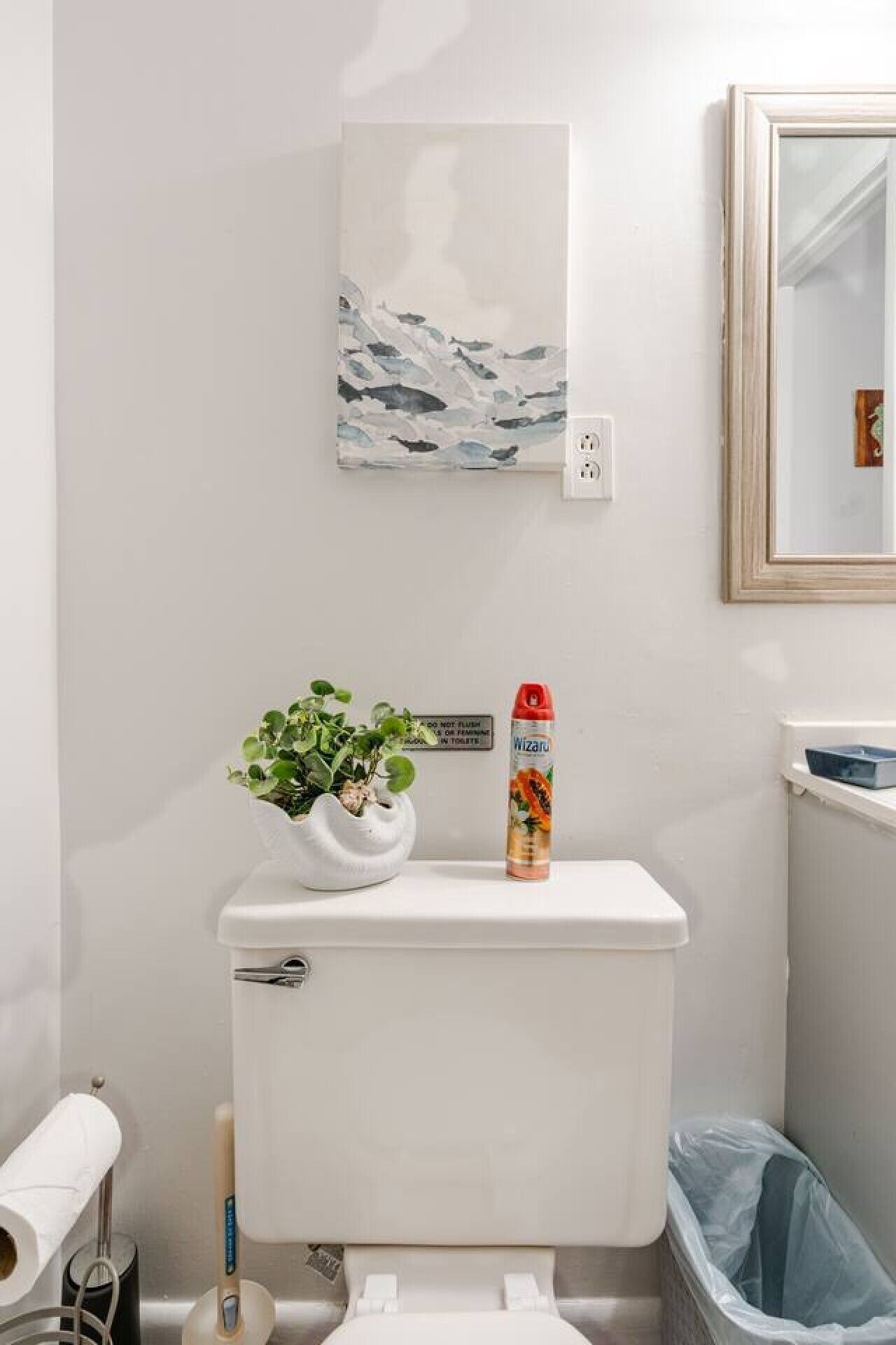 Toilet med hvid tank, et ophængt billede af fisk, en potteplante og toiletpapirholder.