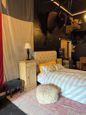 Ein Schlafzimmer mit einer gesteppten Kopfstütze, einem Bett mit gestreifter Bettwäsche, einer Bank, einer Lampe und einem wandmontierten Spiegel.