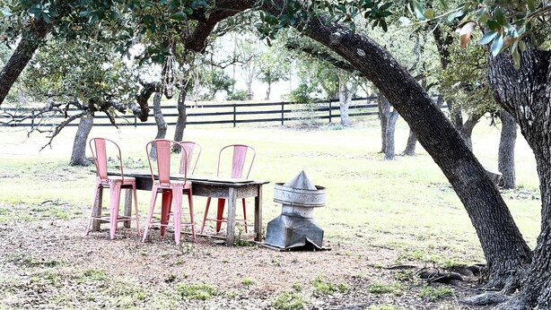 An outdoor seating area with a table and chairs under a tree, a fire pit, and a fenced area in the background. - Hill Country Estate on 11 Acres near Dripping Springs and Wimberley (Wimberley)