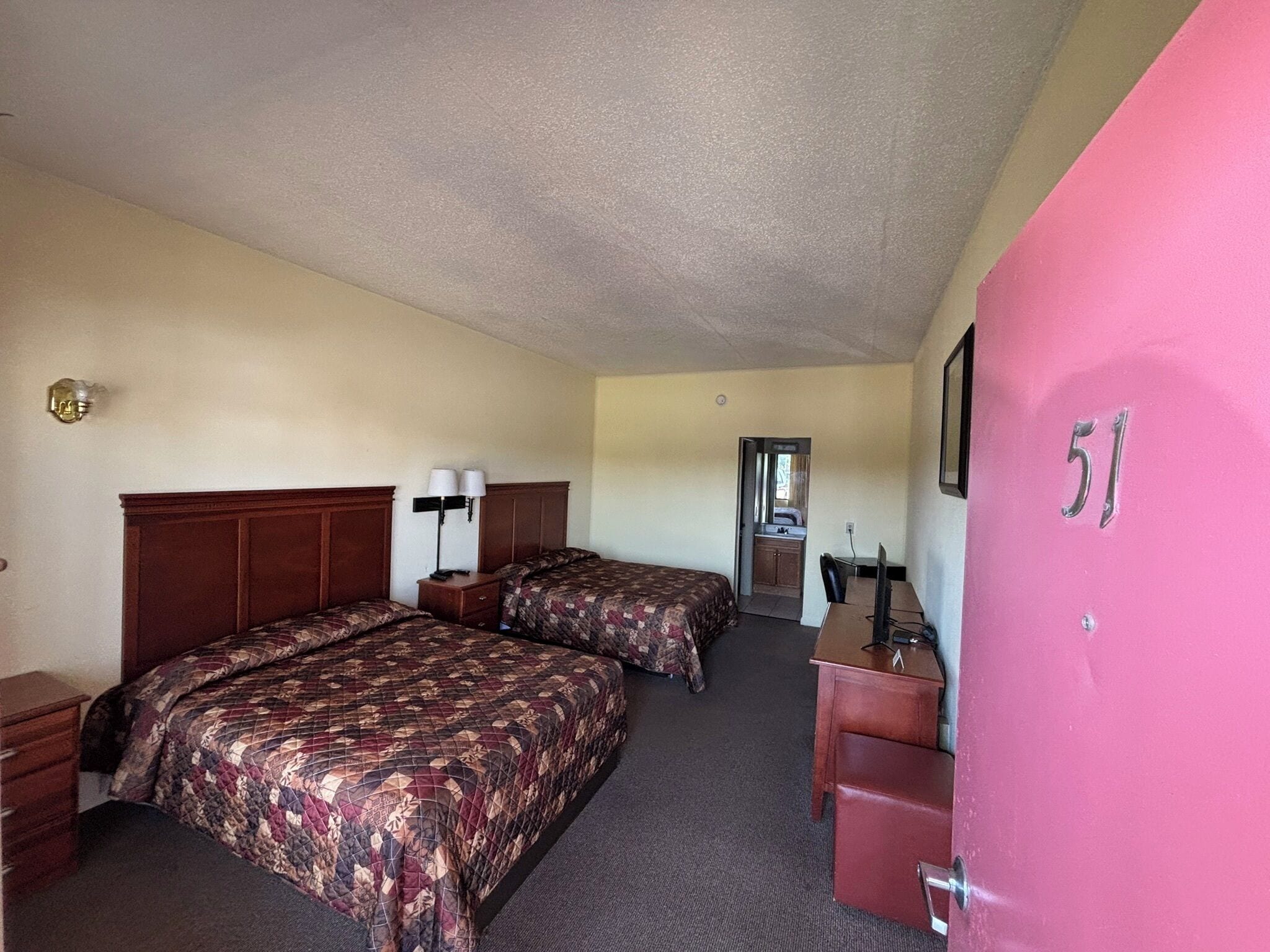 A hotel room with two beds, a desk, a chair, a dresser, and a bathroom visible in the background.