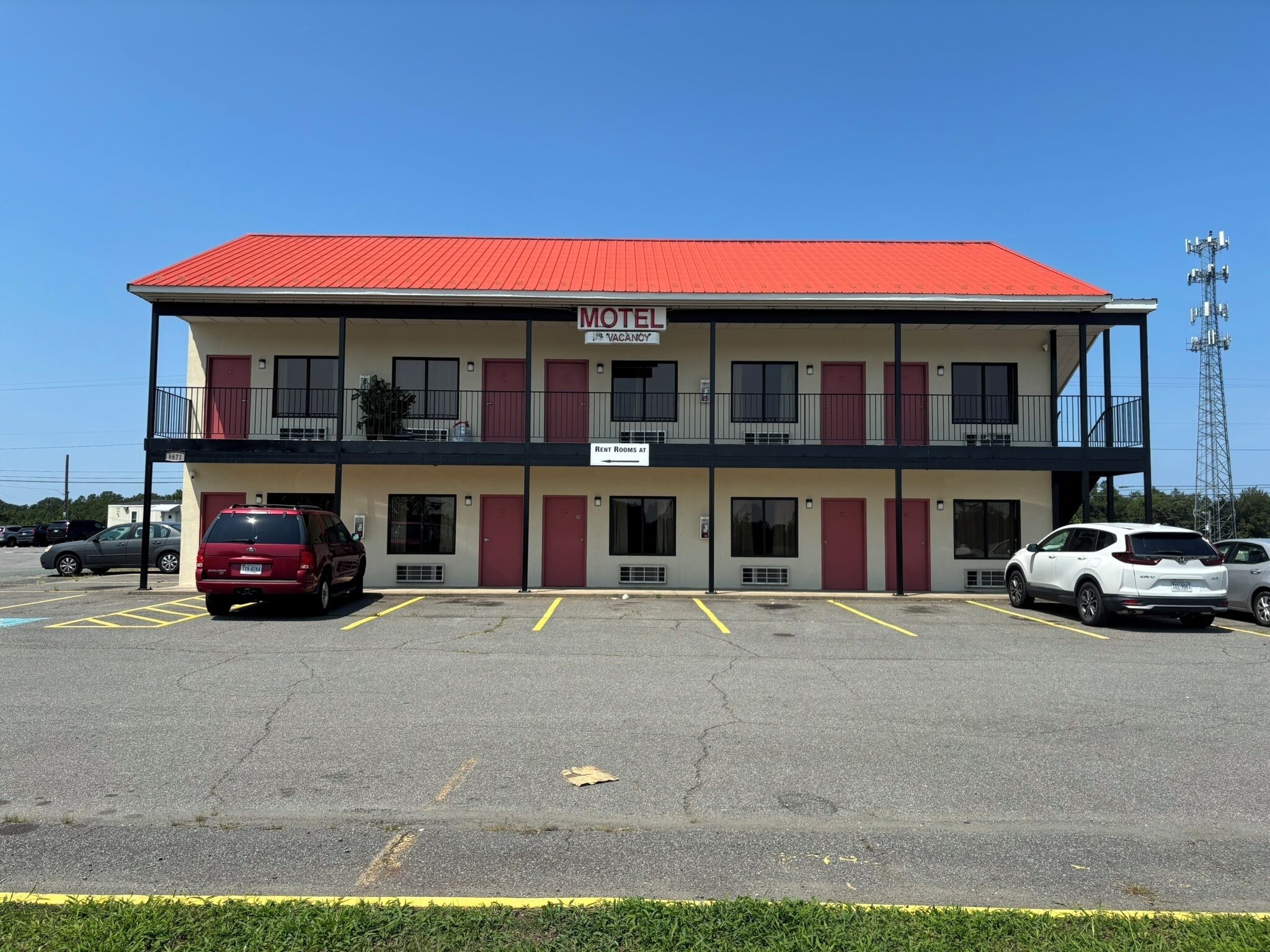 A motel with a red roof, multiple rooms, and a parking lot with several cars.