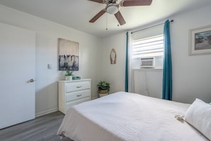 A bedroom with a bed, a dresser, a ceiling fan, a window with curtains, and a wall-mounted air conditioner. - Ocean Breeze 2BR Apt by Wavepool (Virginia Beach)